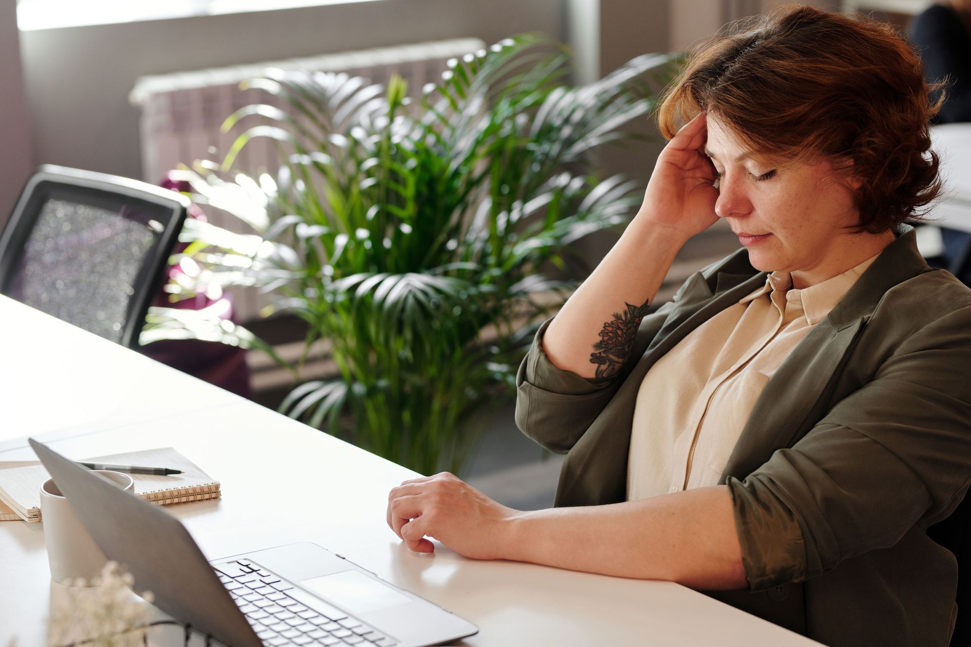 Person sitting at a desk, holding their head while looking at a laptop in a bright office