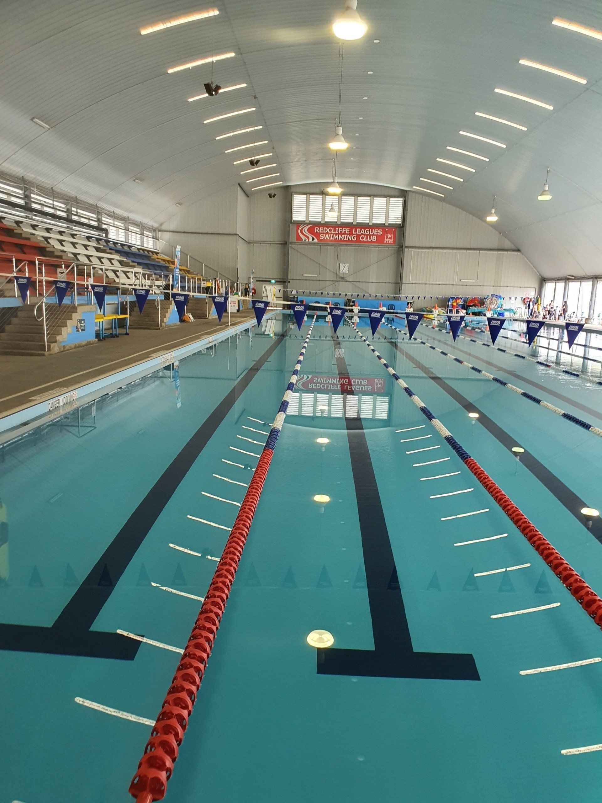 Indoor swimming pool with multiple lanes, swimmers, and spectators under a high arched ceiling.