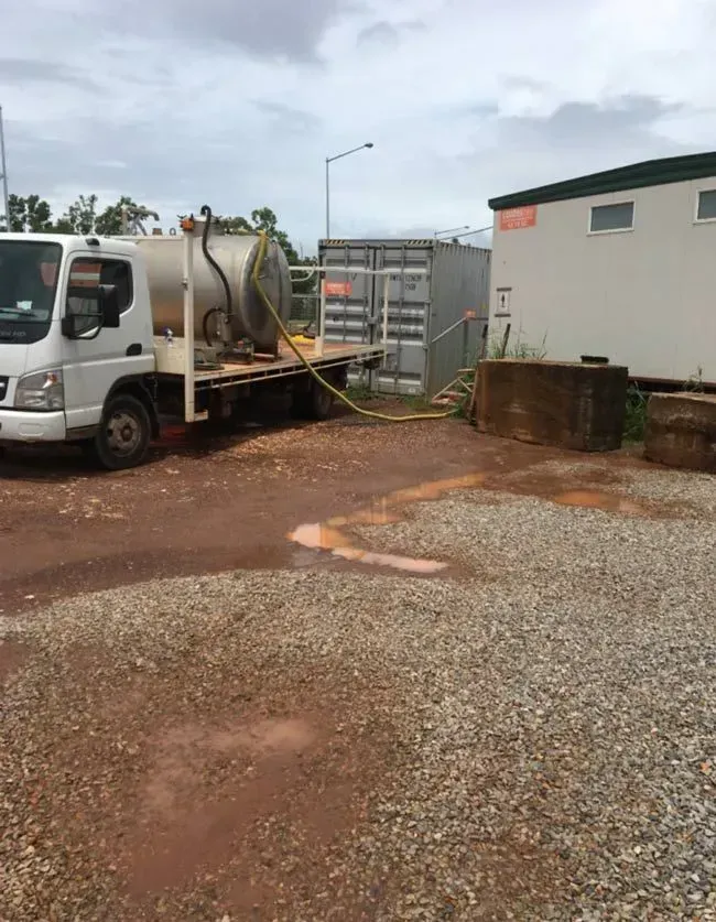 White Utility Truck and Hose Beside a Building on a Muddy Gravel Lot Under Cloudy Skies — Darwin Toilet Hire in Rosebery, NT
