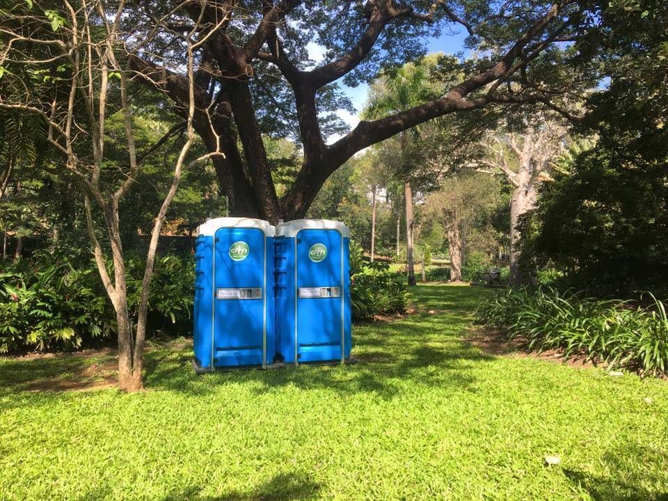 Two Blue Portable Toilets Standing on a Grassy Park Lawn Surrounded by Trees — Darwin Toilet Hire in Rosebery, NT
