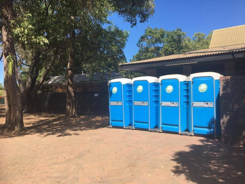 Four Blue Portable Toilets Lined Up Outdoors Beside a Building and Trees — Darwin Toilet Hire in Rosebery, NT