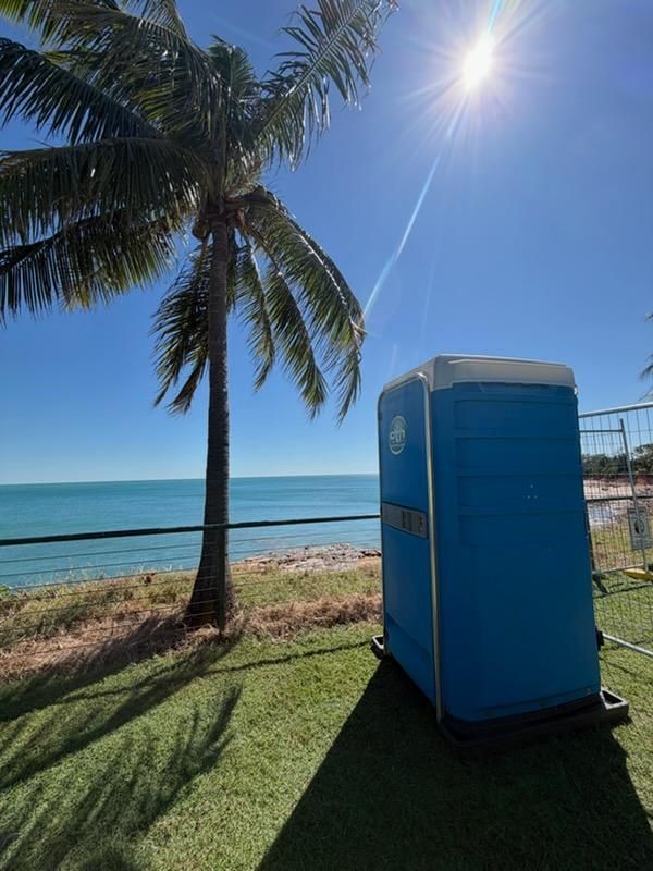 Blue Portable Toilet on Grassy Beach by Palm Tree and Ocean Under Bright Sun — Darwin Toilet Hire in Katherine, NT