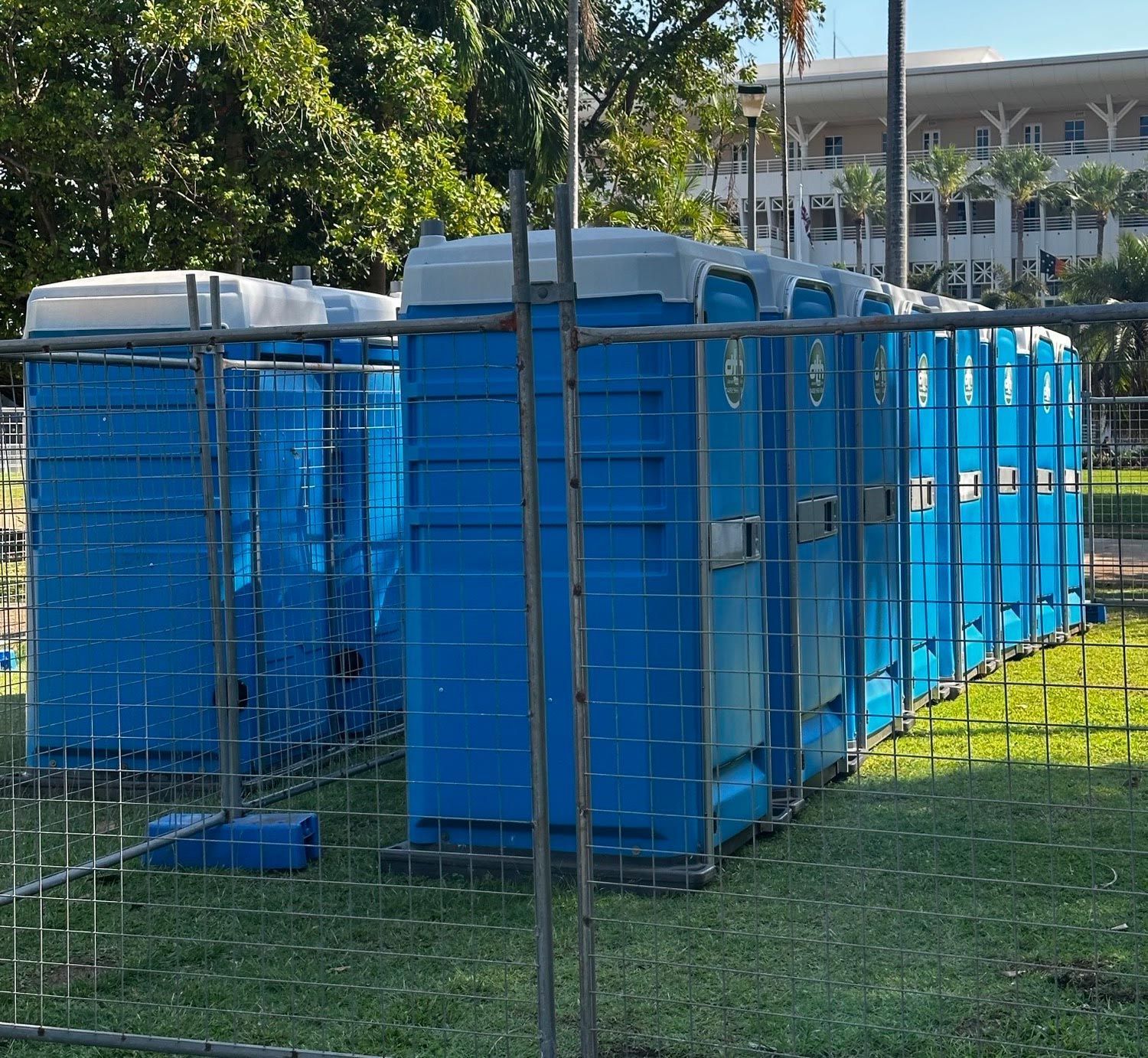 Blue Portable Toilets Lined Up Behind a Fence on a Grassy Outdoor Area — Darwin Toilet Hire in Rosebery, NT