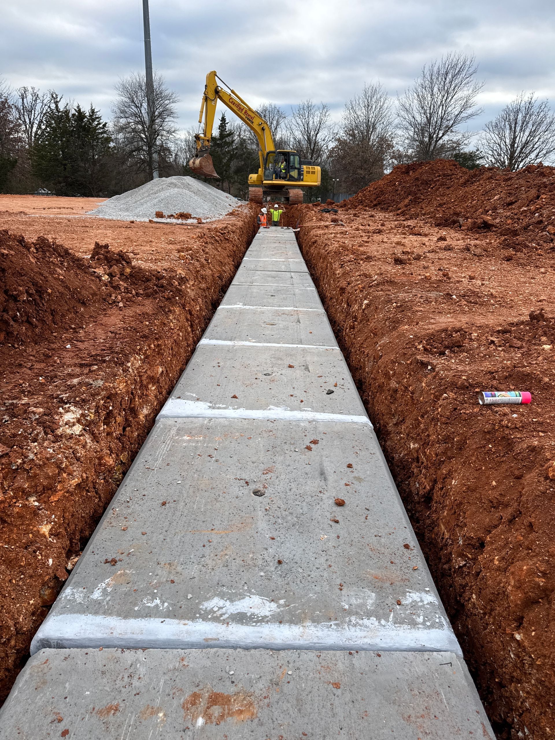 Excavator working on a concrete pathway between earthen berms, construction site.