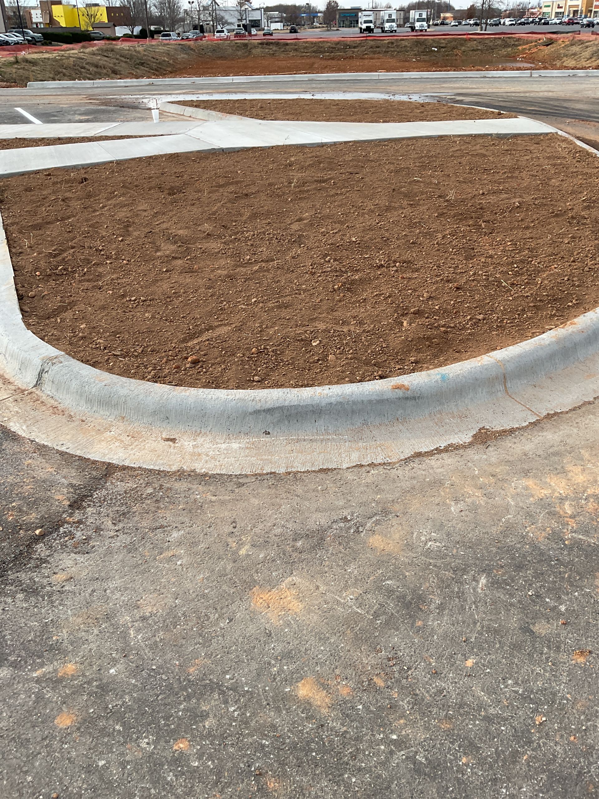 Brown mulch in a curved concrete planter in a parking lot, with asphalt in the foreground.