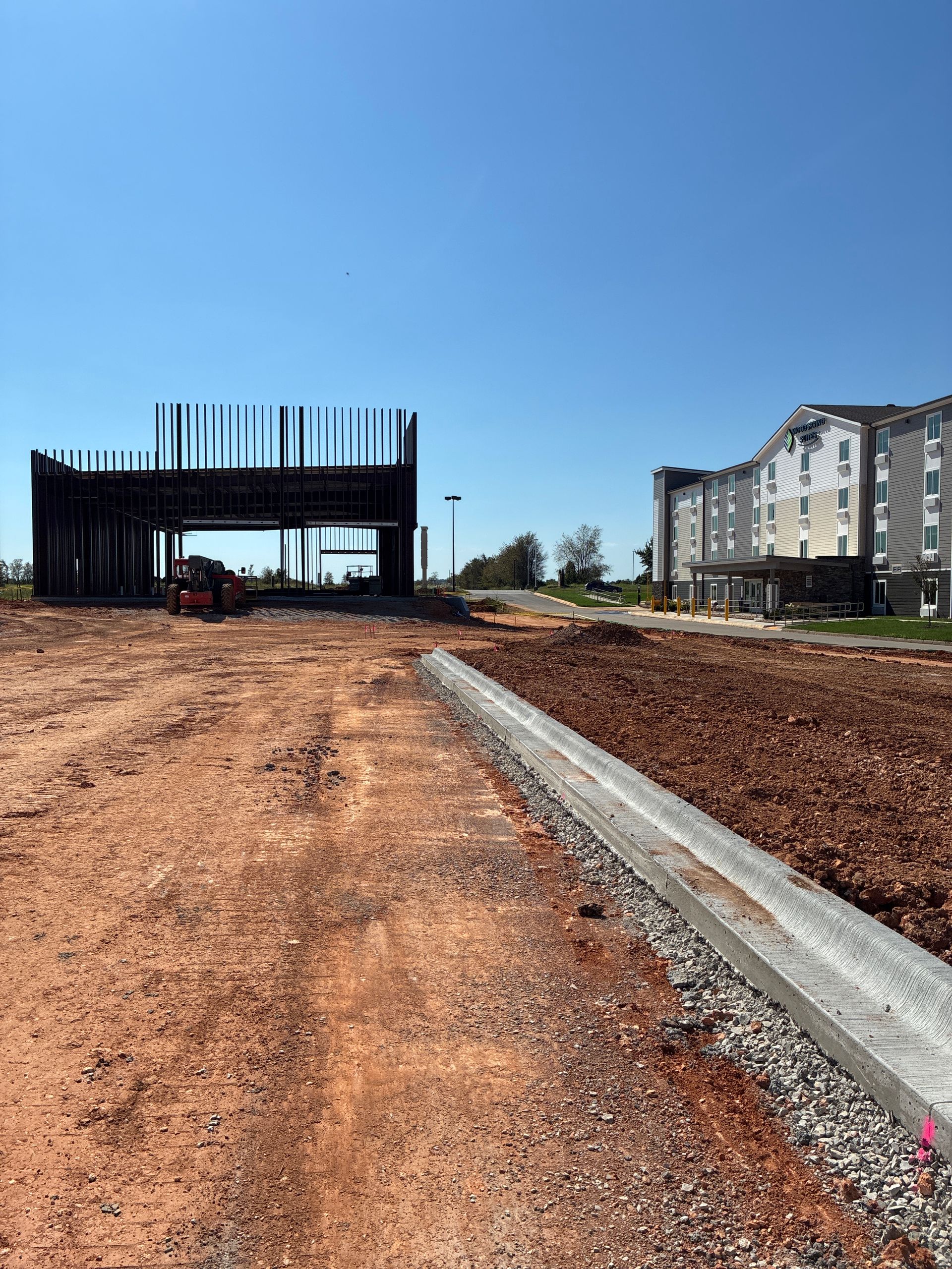 Construction site with unfinished building frame. Dirt, gravel, concrete curb, and a multi-story building in the distance. Blue sky.
