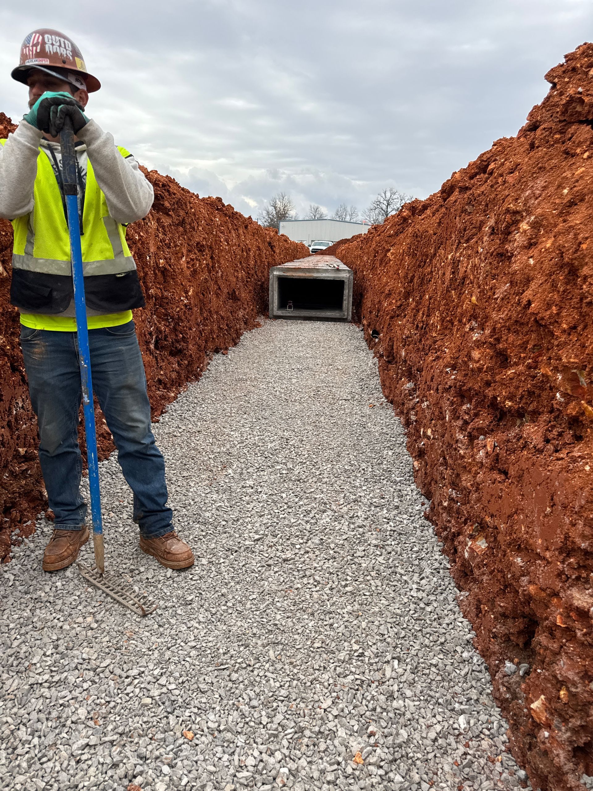 Construction worker stands in a gravel-filled trench, holding a shovel; a concrete culvert is in the distance.