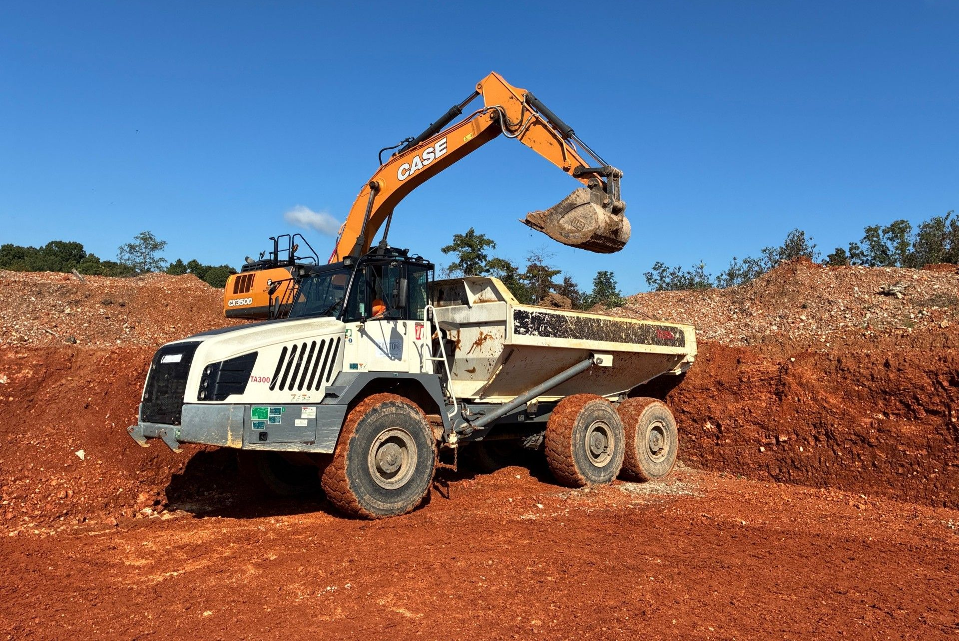 An excavator loading a dump truck with red earth at a construction site against a blue sky.