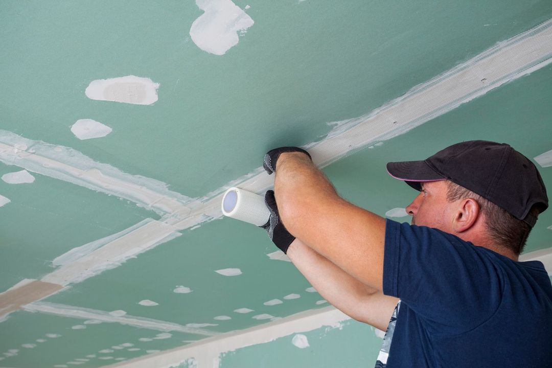 Persona che installa nastro adesivo per cartongesso su un soffitto verde, indossando un berretto nero e guanti.