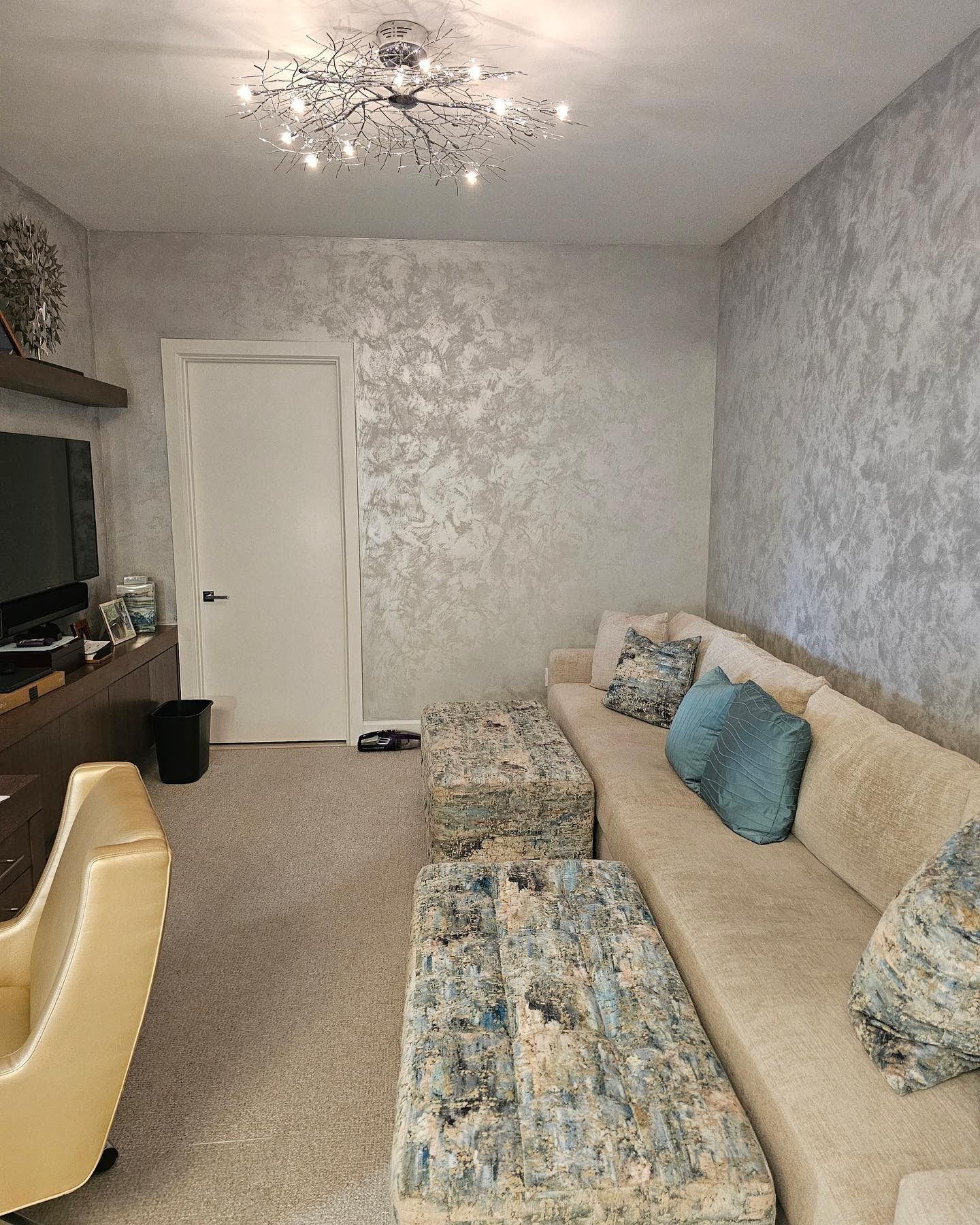 Living room with textured silver walls, beige sectional, patterned ottomans, and a bright ceiling light.
