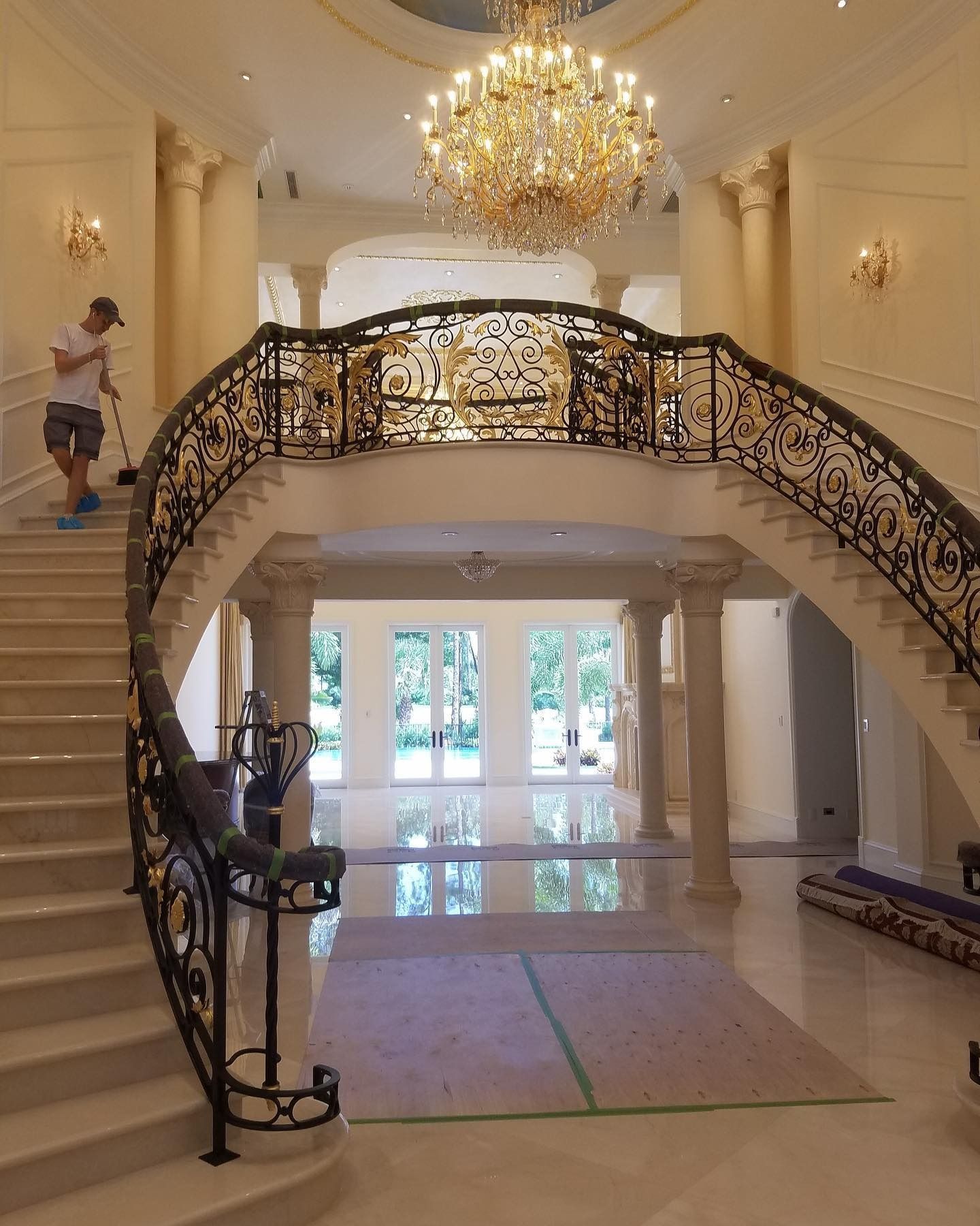 Man sweeping grand foyer with ornate staircase, chandelier, columns, and large glass doors.