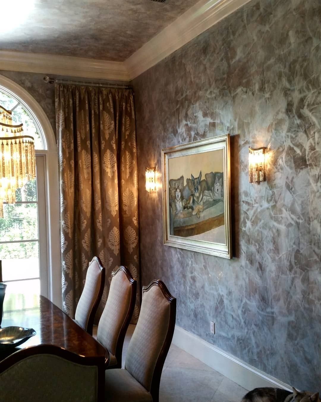 Dining room with ornate chairs, a dark wooden table, textured gray walls, gold framed artwork, and a chandelier.