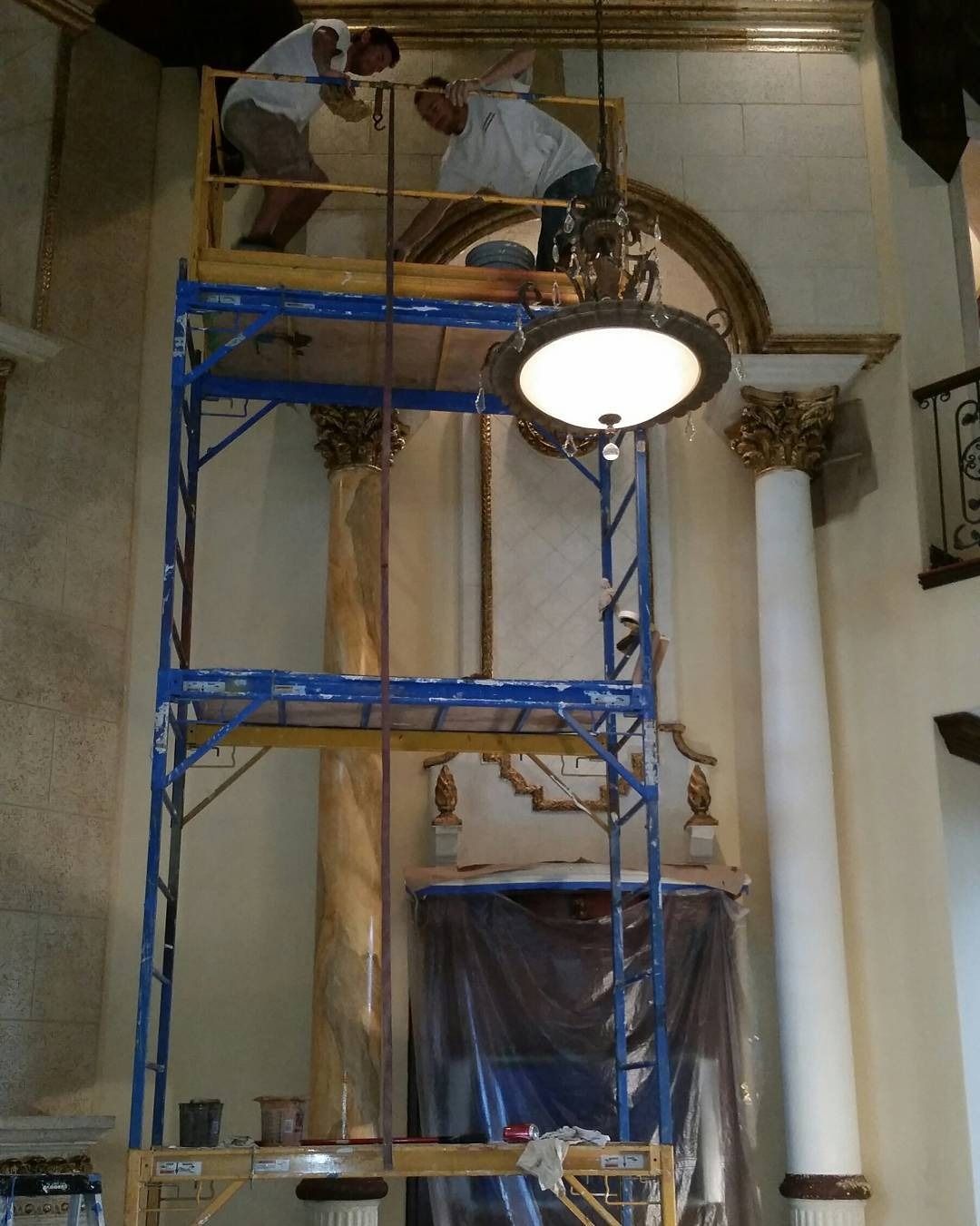 Two workers on a blue scaffolding, working on a wall with columns and ornate gold trim, inside.
