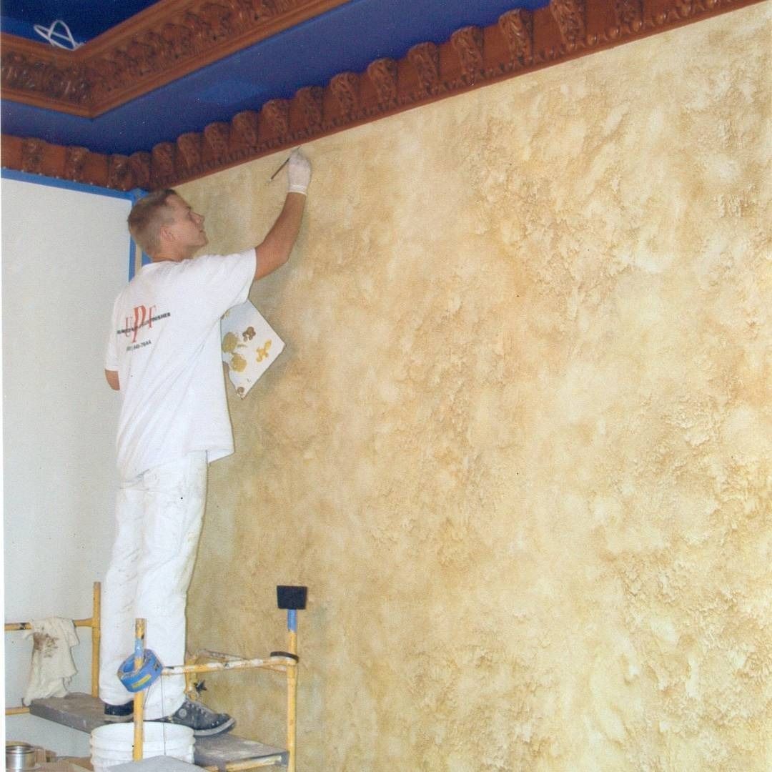 Painter on a scaffold applying faux finish to a beige wall. Blue ceiling with decorative trim.