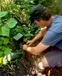 A man is kneeling down in the woods working on a device.