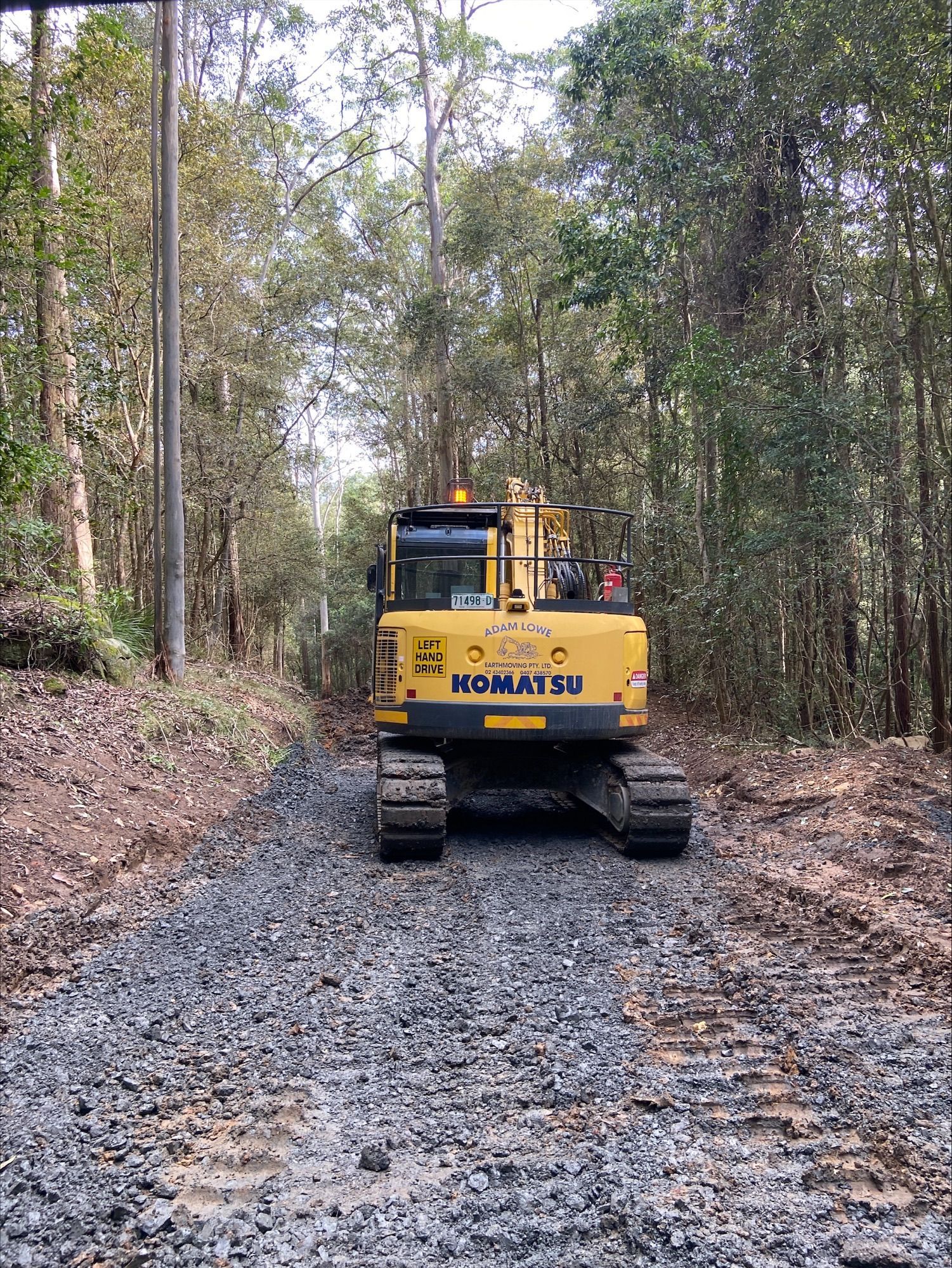 A yellow excavator is driving down a dirt road in the woods. — Adam Lowe Earthmoving Pty Ltd in Somersby, NSW