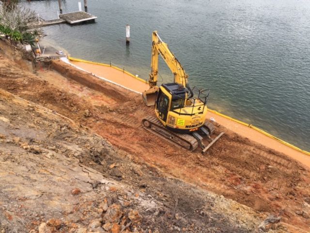A yellow excavator is driving down a dirt road next to a body of water. — Adam Lowe Earthmoving Pty Ltd in Somersby, NSW