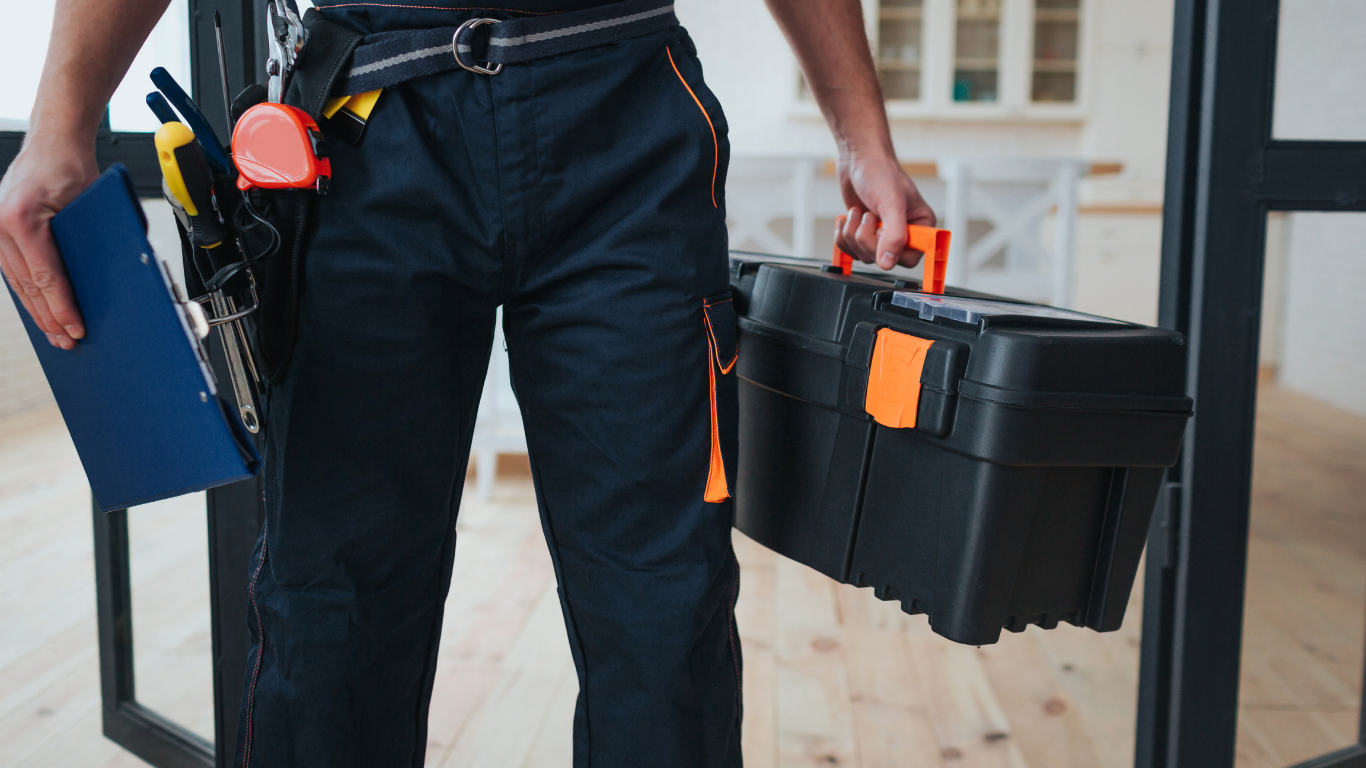 Worker carrying a black toolbox and clipboard, wearing dark work clothes and safety gear.