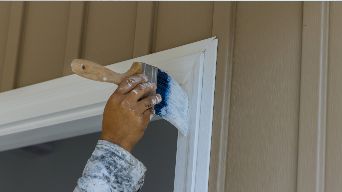 Person painting white trim with a brush on beige siding