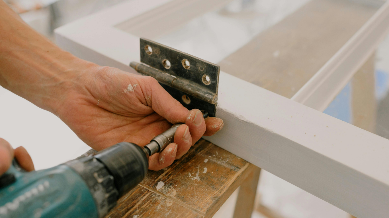 Hands using a drill to attach a black bracket to a white wooden frame.