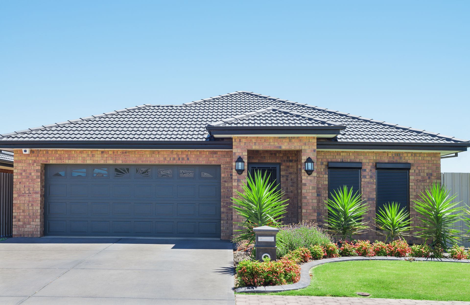 Photograph showing a single-story suburban brick house with a broad tiled roof, double garage, dark shutters, and front entry.