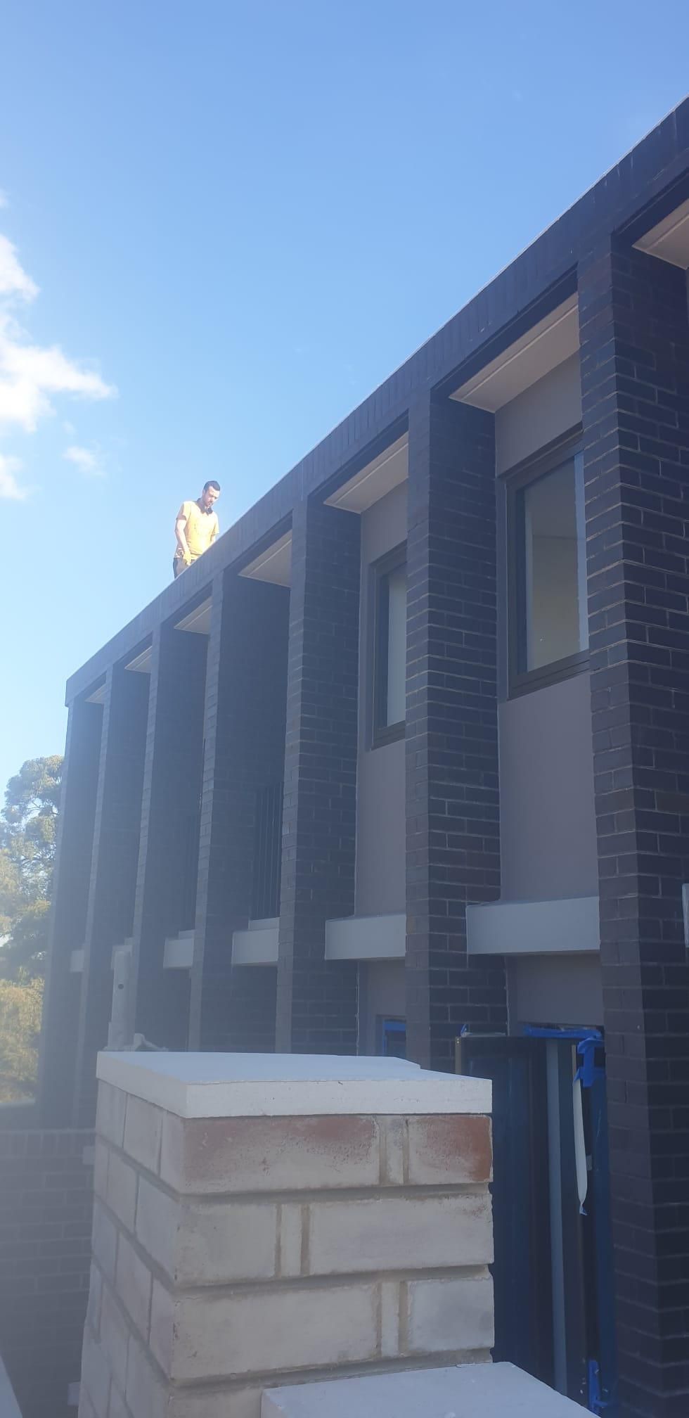Construction worker in blue helmet and safety harness smiling, arms crossed in front of brick construction site.