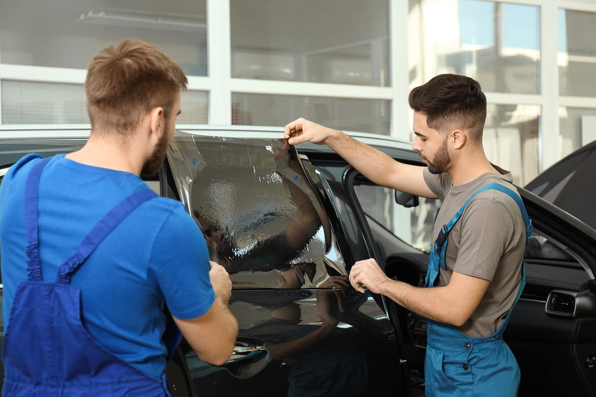 Two men are installing window tinting on a car in a garage.