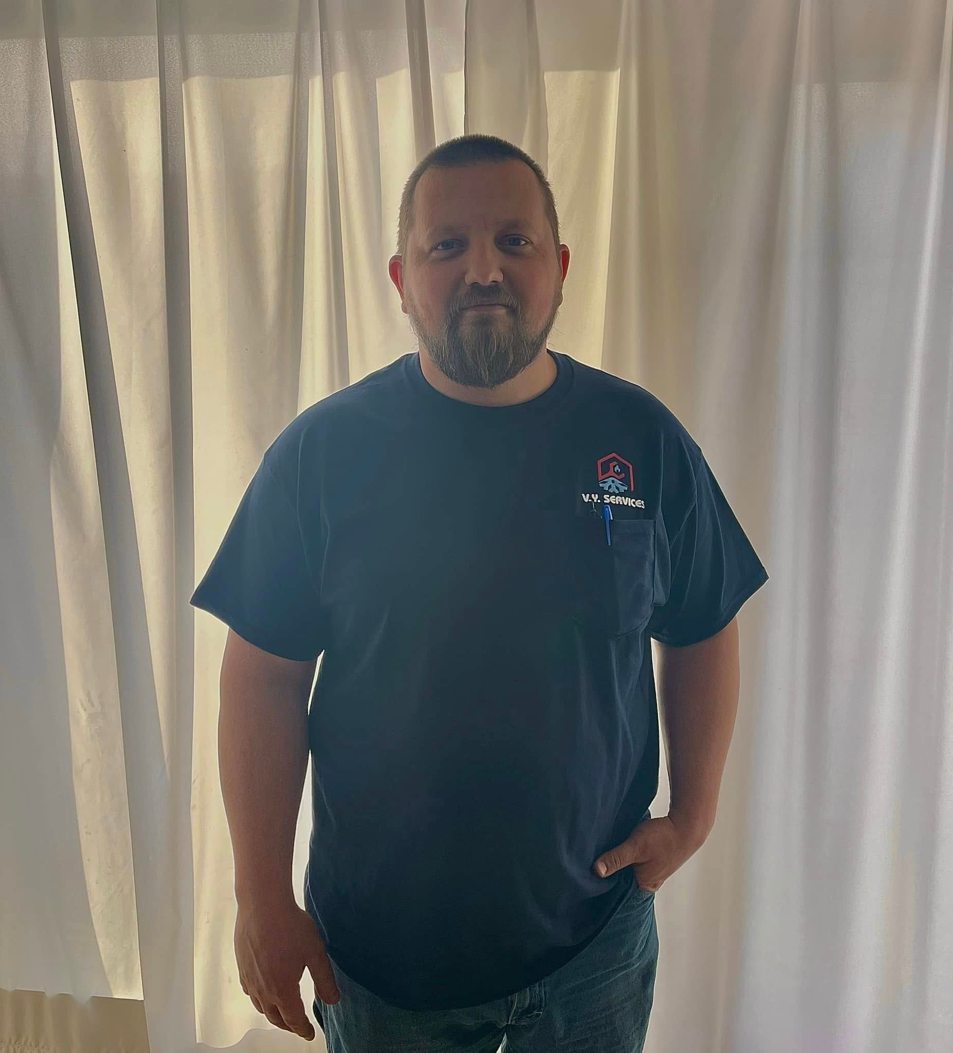 A man with a beard wearing a black t-shirt is standing in front of a window.