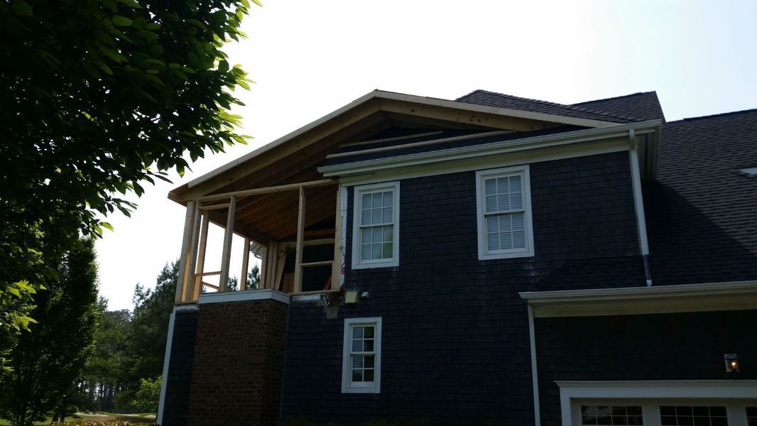 Construction on a two-story house, a framed porch addition. Dark blue siding, brick base, white trim, trees.