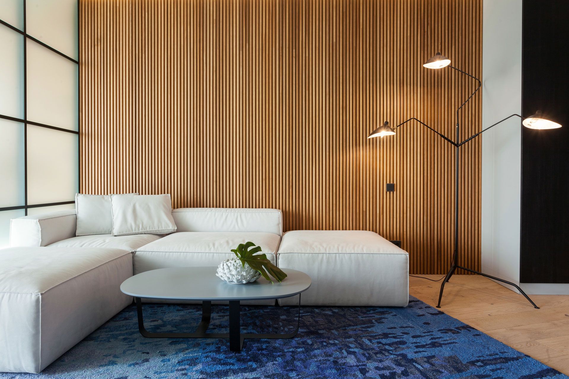 Modern living room with white sectional sofa, blue rug, wooden wall, and black floor lamp.