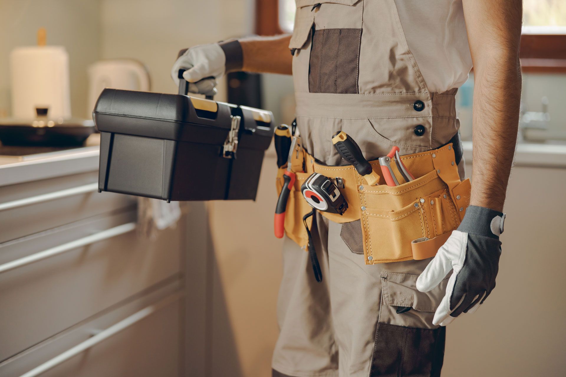 Person in work overalls, tool belt, and gloves holding a black toolbox indoors.