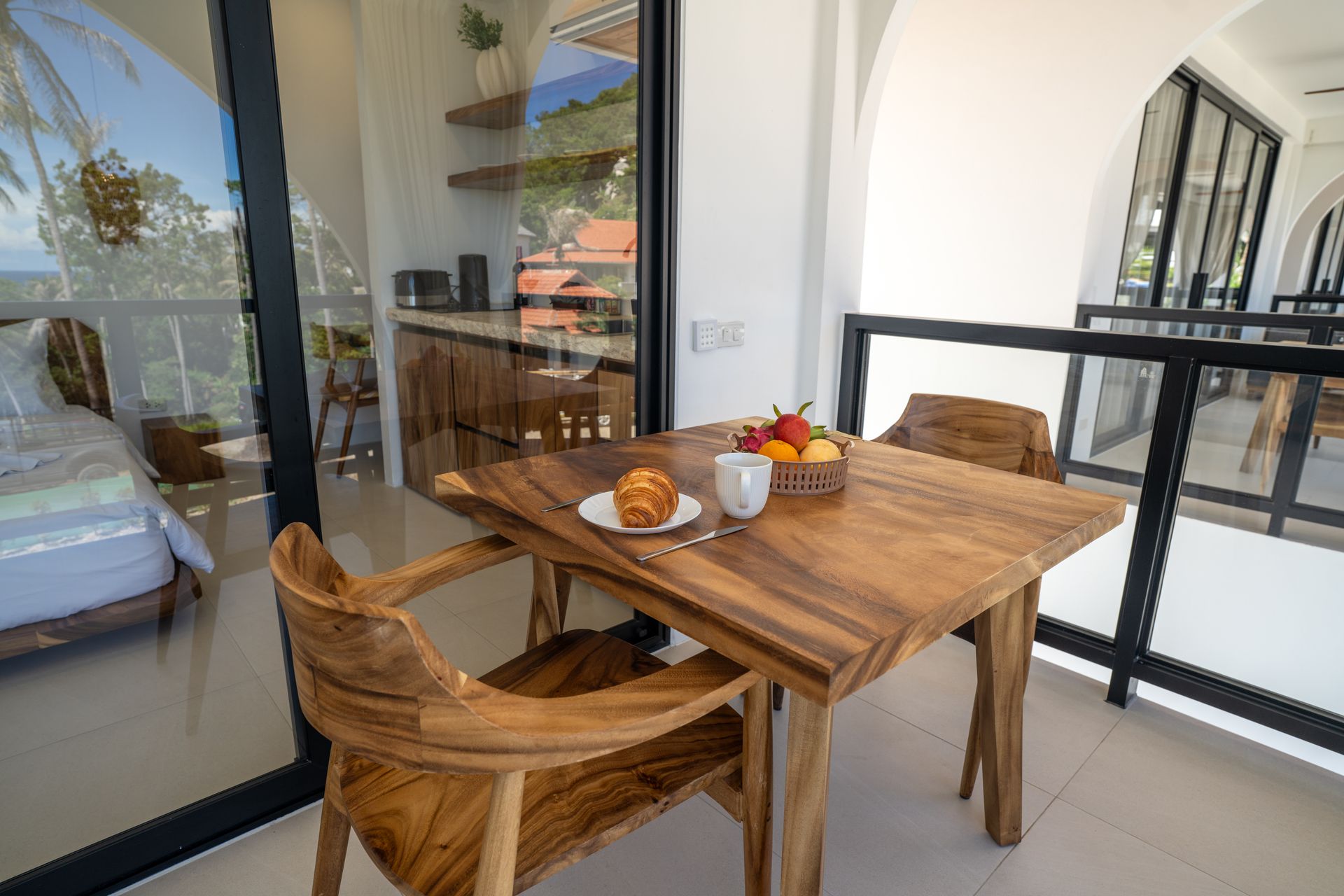 Wooden dining table and chairs on a balcony with fruit, coffee cups, and glass railing view