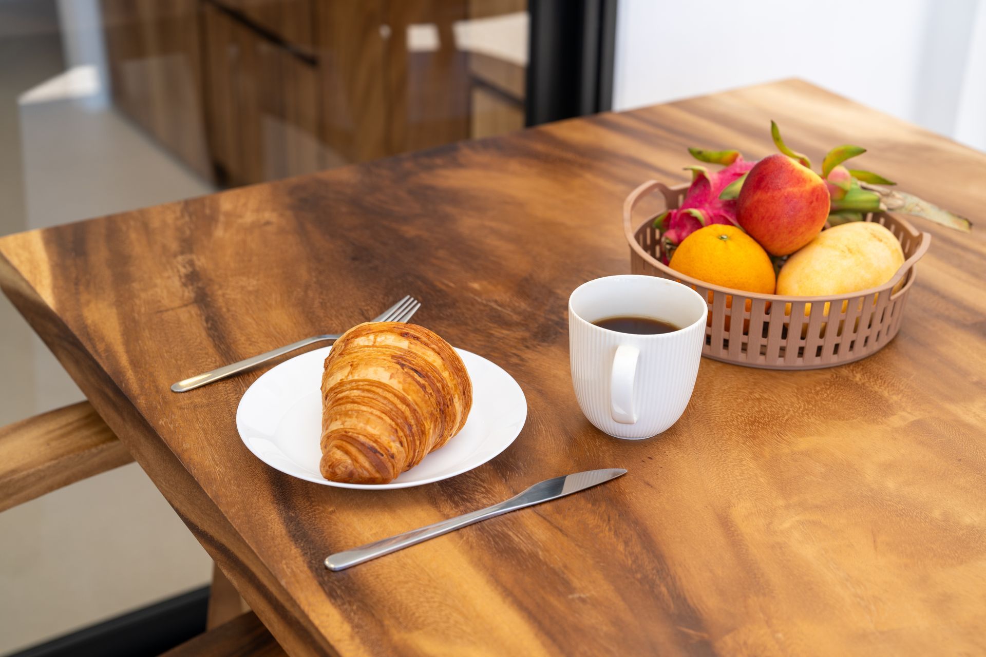 Croissant, coffee mug, and fruit basket on a wooden table near a window.