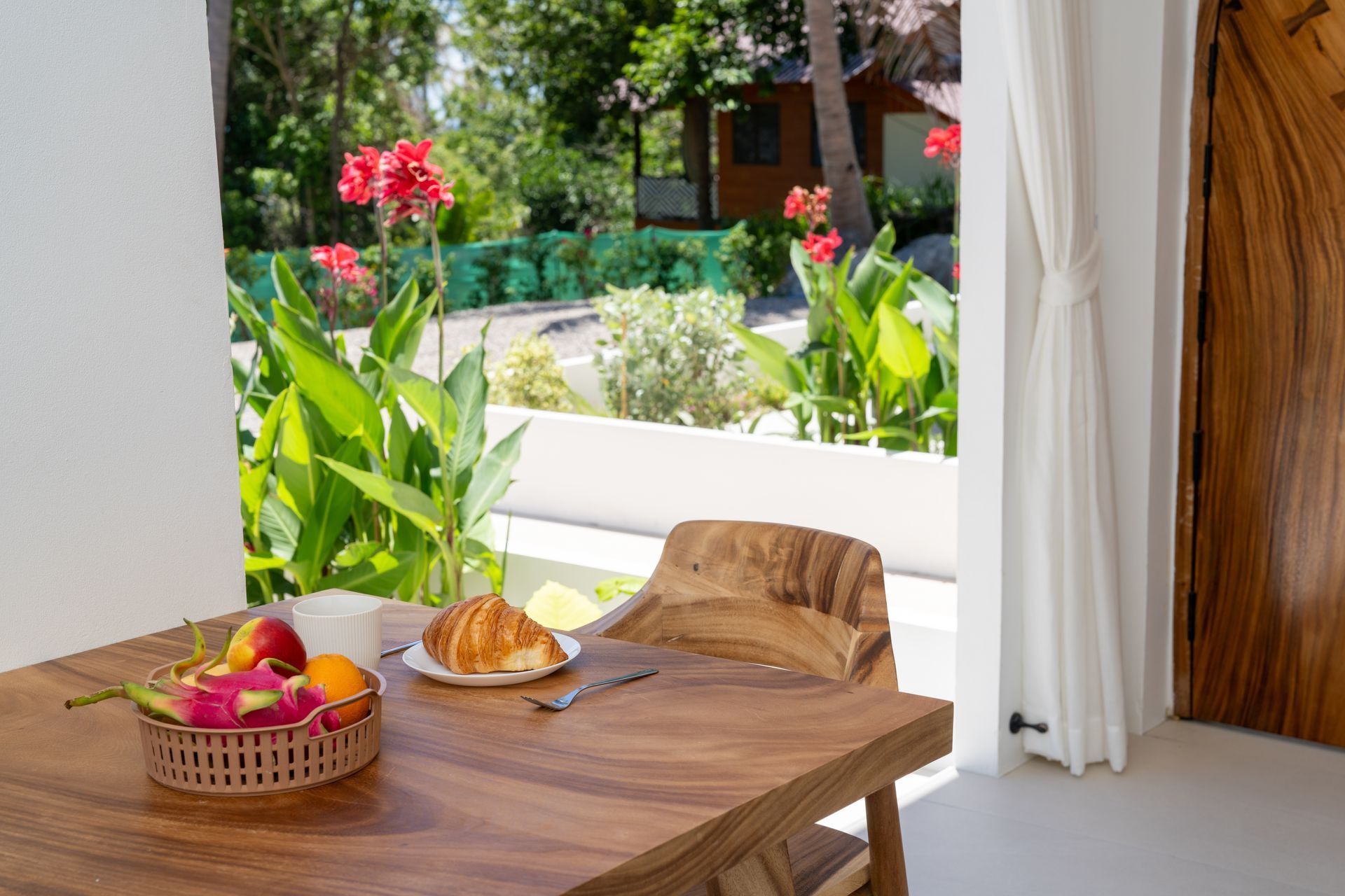 Wooden table with fruit and croissant by an open window overlooking a lush garden