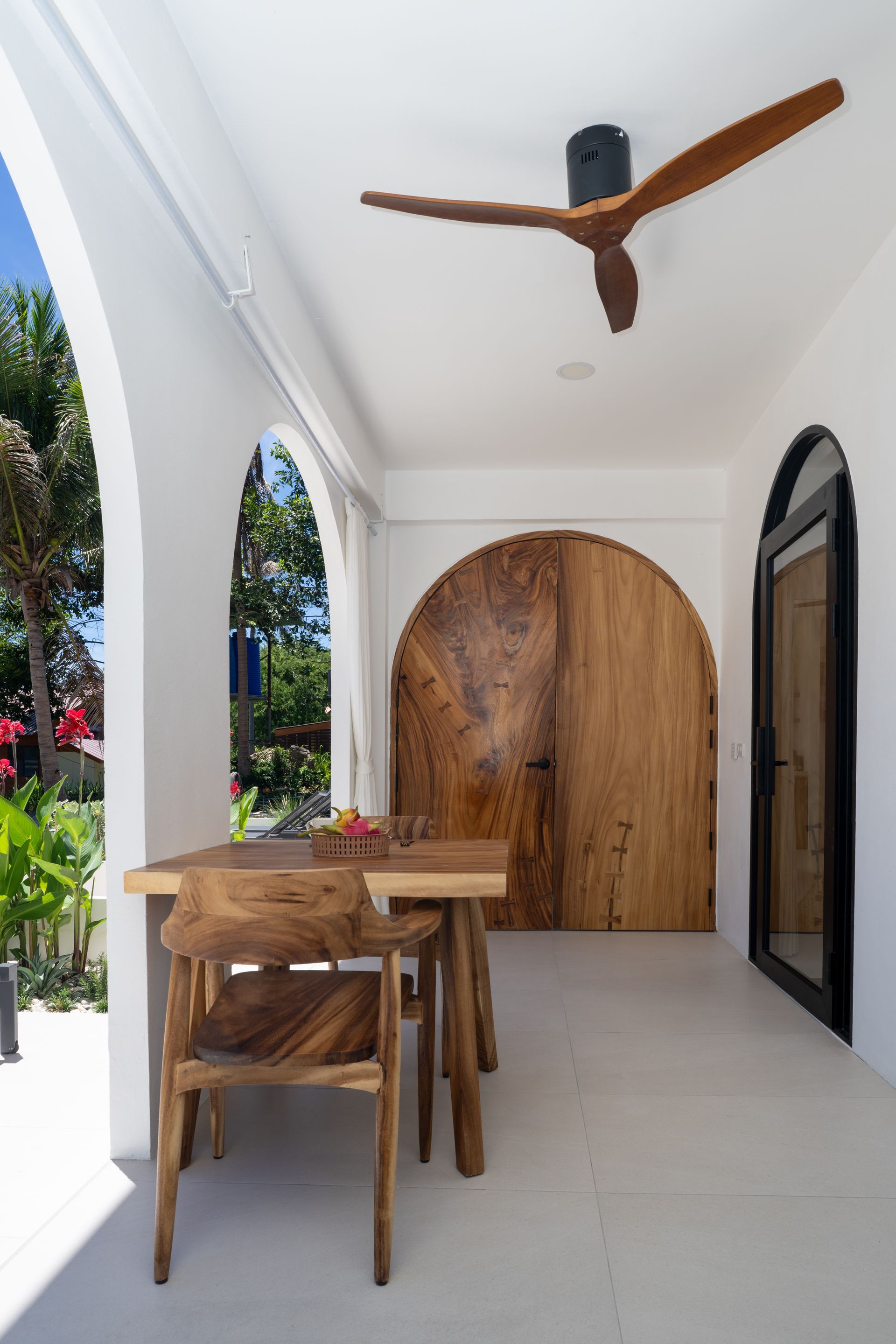 Bright white patio with wooden table, chairs, arched doorway, and ceiling fan.