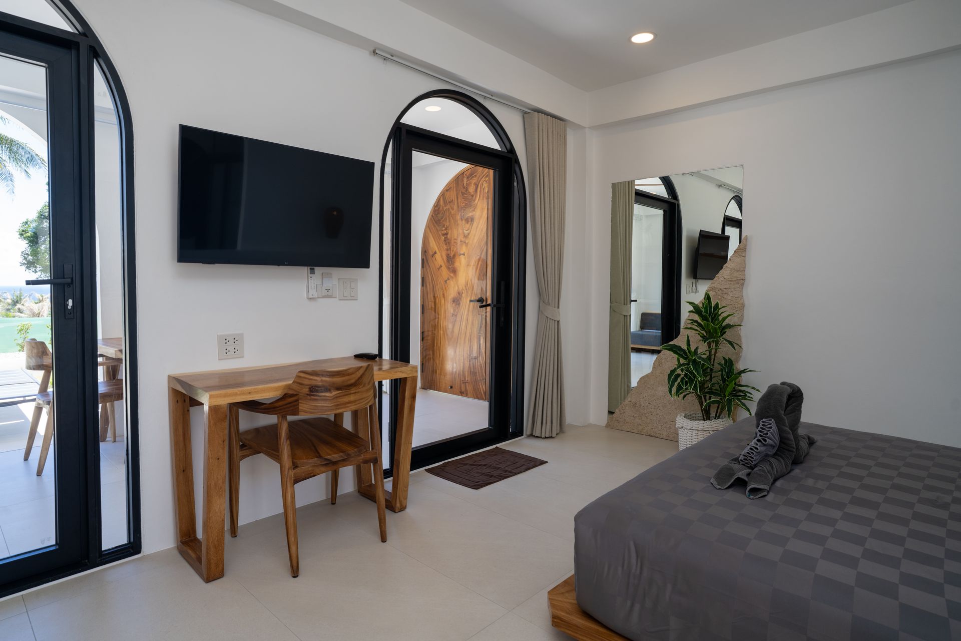 Bright bedroom with wall-mounted TV, small desk, arched door, and bed by curtains and a potted plant