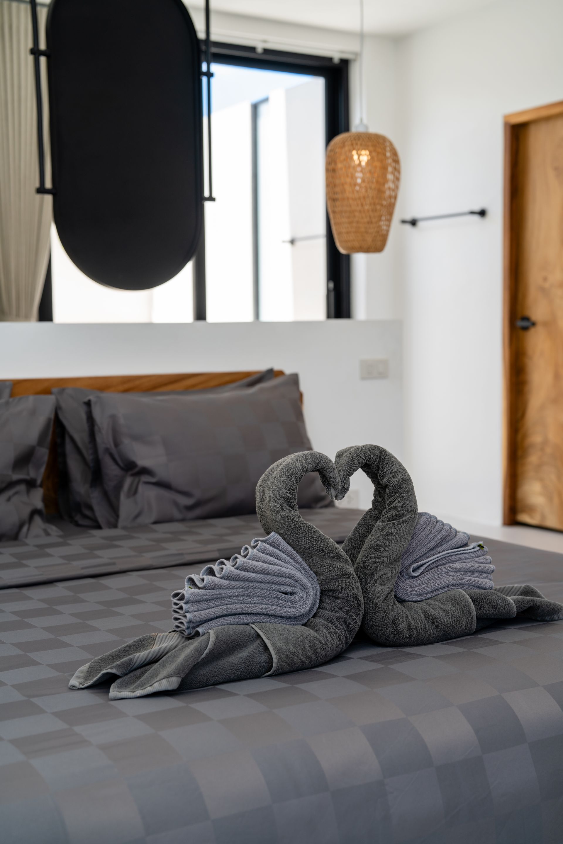 Gray bed with swan-shaped towel art in a modern bedroom with black curtains and wicker pendant lamps.