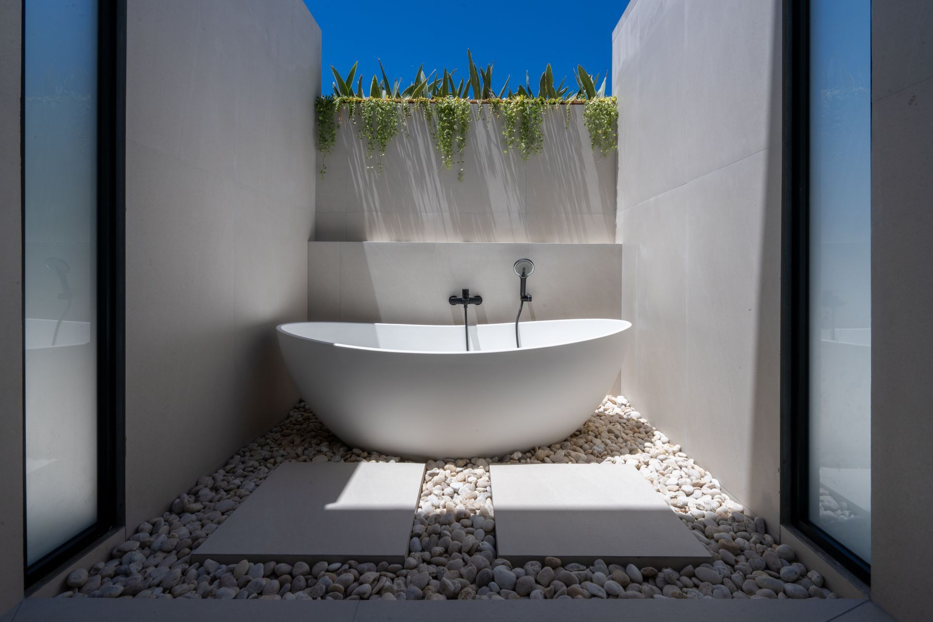Minimalist outdoor bathtub in a white pebble courtyard, framed by white walls and a blue sky.