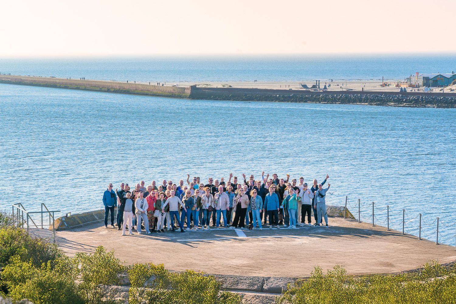 Een groep mensen zwaait op een betonnen platform bij de zee, met een golfbreker op de achtergrond.