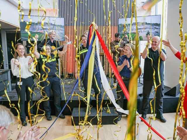 Een groep mensen viert feest en knipt linten door met slingers. Binnen, sommigen in uniform.