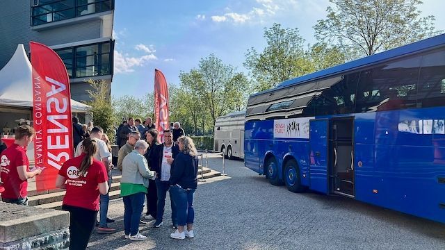 Mensen verzamelen zich bij een blauwe bus en een gebouw met rode banners.