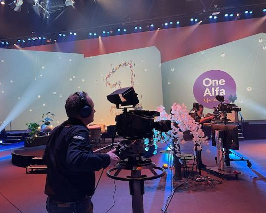 Iemand bedient een televisiecamera op een toneeldecor met het logo 