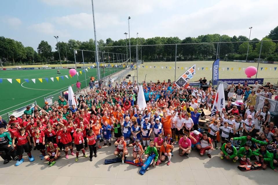 Grote menigte in kleurrijke sportuniformen bij een hockeywedstrijd. Groen veld, blauwe lucht.