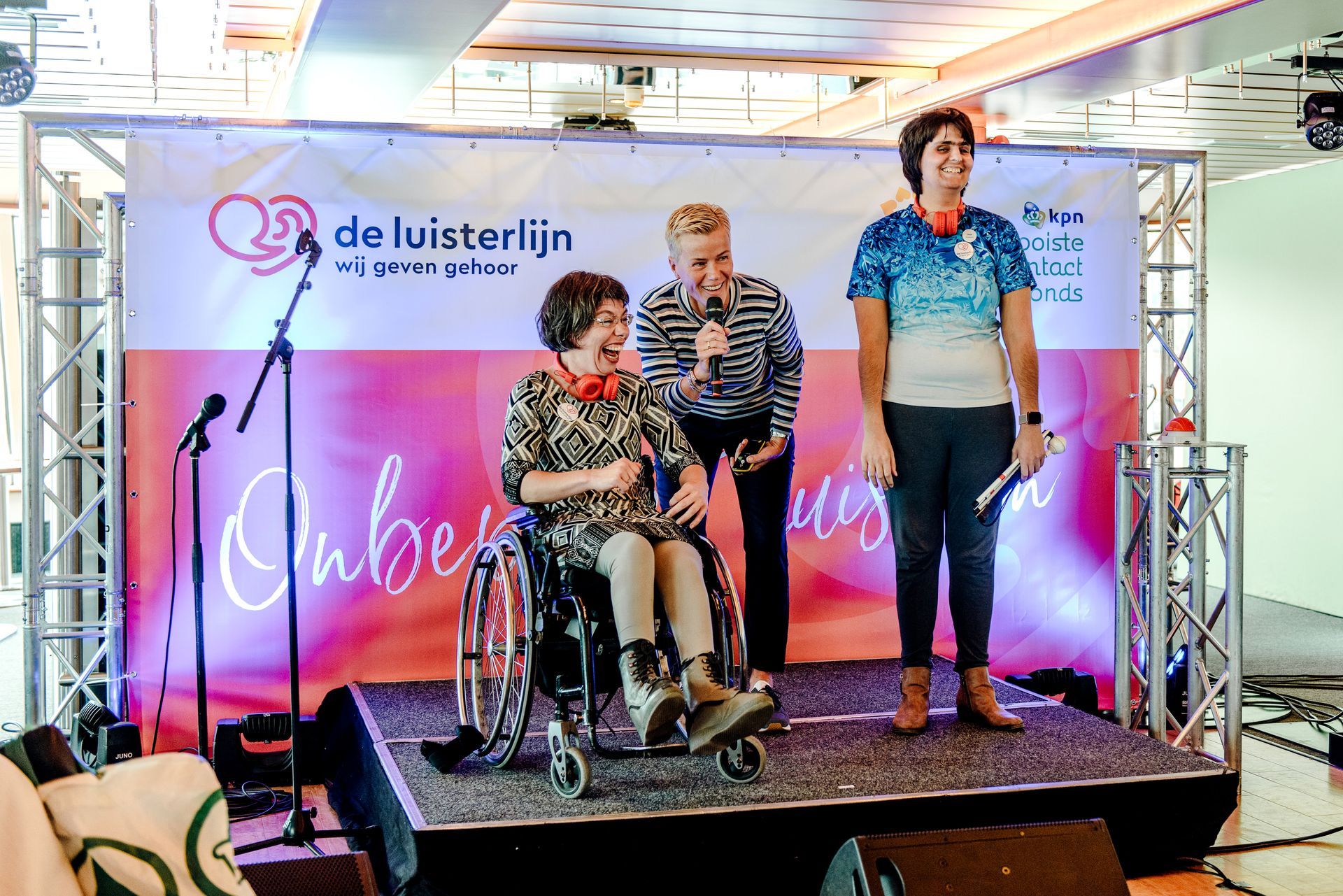 Drie mensen op het podium, waarvan één in een rolstoel, met microfoon in de hand voor het spandoek 