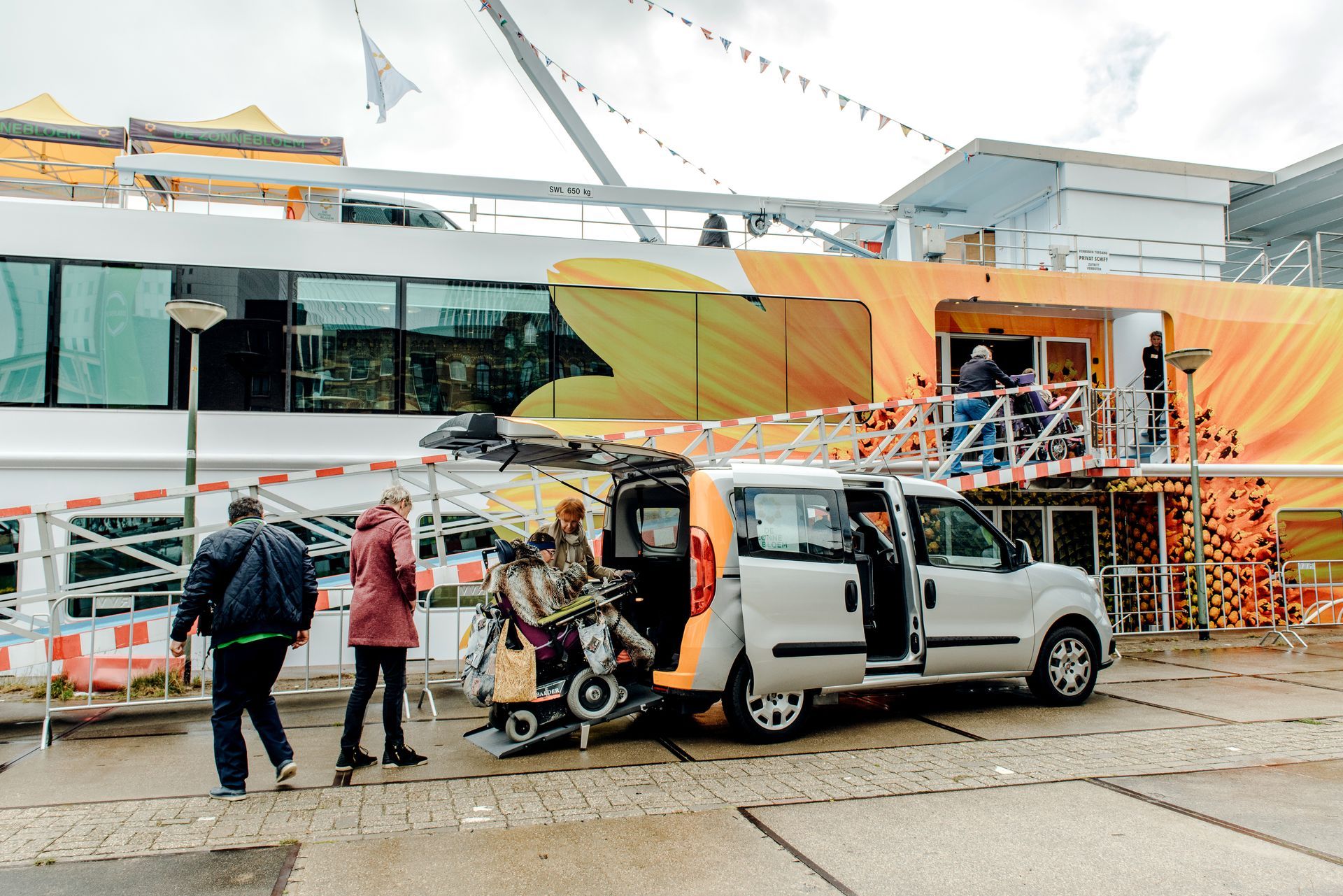Een rolstoelbusje laadt een rolstoel uit bij een gebouw met een oranje en geel ontwerp. Mensen staan ​​ernaast.