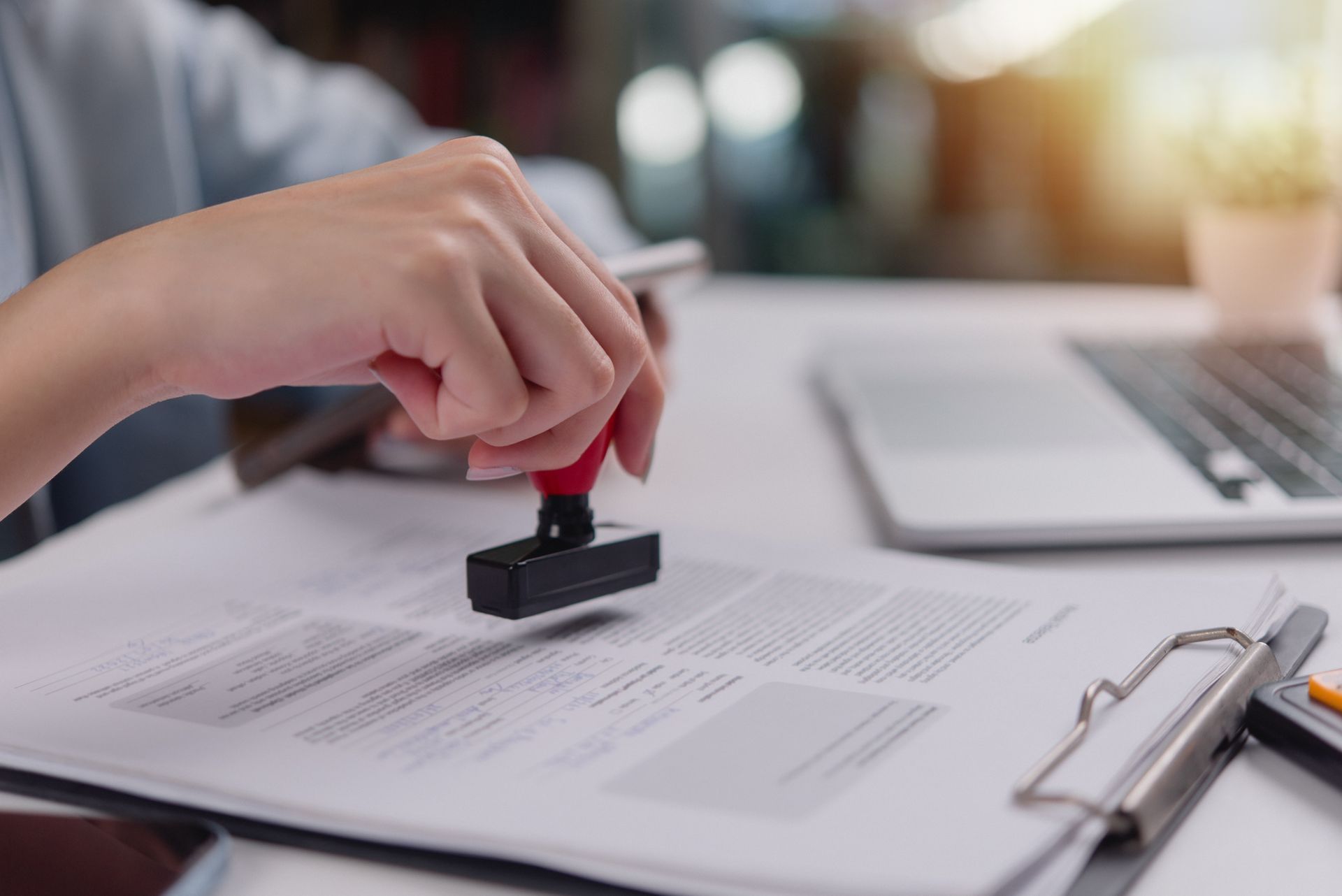 Hand stamping a document with a red stamp, office setting with laptop and clipboard.