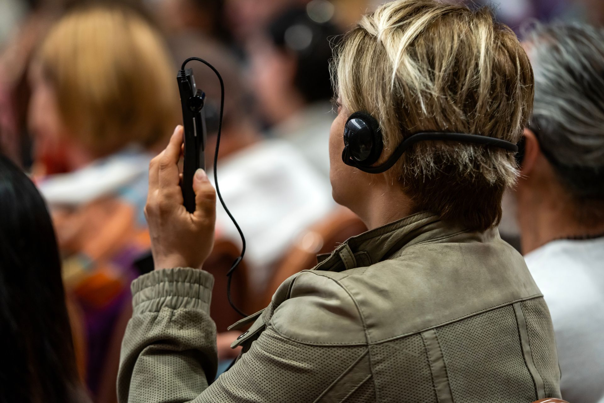 Woman in tan jacket, using a headset and handheld device, likely for interpretation or audio guide, in a crowded indoor setting.