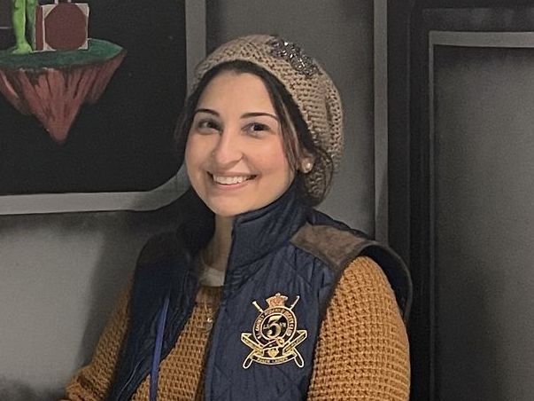 Woman smiling, wearing a knitted cap, vest, and sweater. Standing indoors with art on the wall.