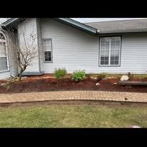 Mulched flower bed with small shrubs along the front of a gray house, beside a curved brick border.