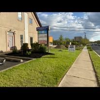 Sidewalk beside beige building and grassy lawn under cloudy sky, with roadside signs and parking lot.